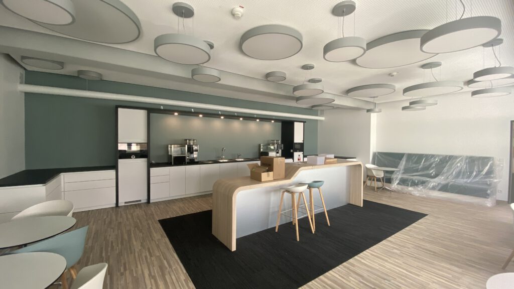 Hochwertige Büroausbau. Eine Küche mit modernen Möbeln, schönen kreisförmigen Lampen und einem mintgrünen Farbschema, hellem Holzton und weißer Front.